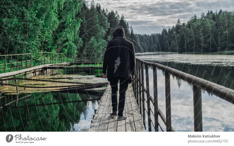 Man in the hood walks along the pier man back alone nature black rear sunny overcast cloud person path journey lonely tree silhouette walking away woods coast