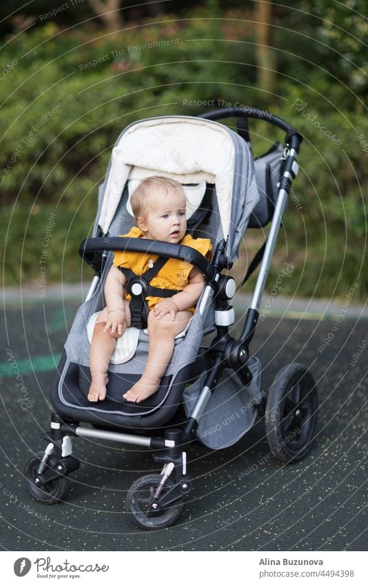 Sweet caucasian baby girl ten months old sitting in stroller outdoors. Little child in pram. Infant kid sits in pushchair. Summer walks with kids. Family leisure with little child.