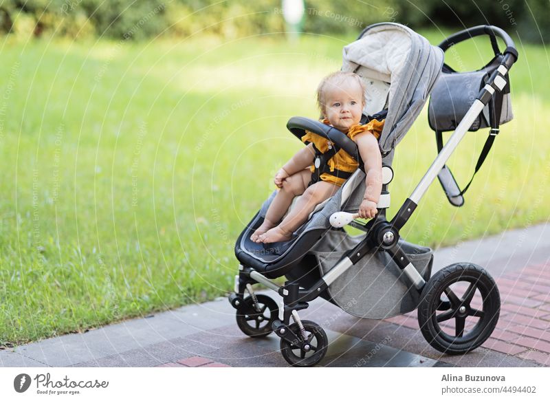 Sweet caucasian baby girl ten months old sitting in stroller outdoors. Little child in pram. Infant kid sits in pushchair. Summer walks with kids. Family leisure with little child.