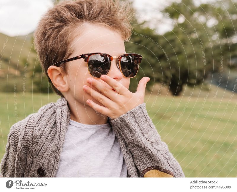 Stylish boy in sunglasses sitting in countryside kid childhood style fashion nature leisure tree field content green trendy carefree summer glad plant verdant