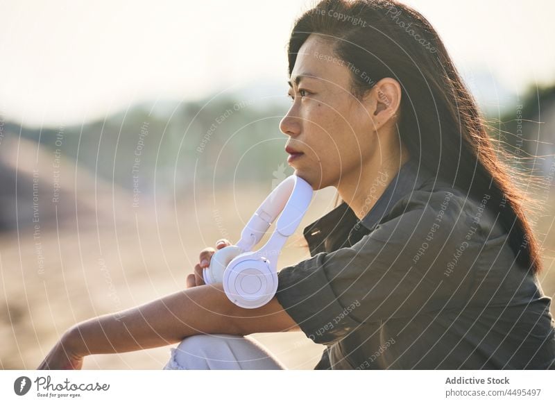 Pensive Asian woman listening to music on beach seaside seashore headphones meloman song pastime thoughtful pensive playlist sand asian female summer recreation