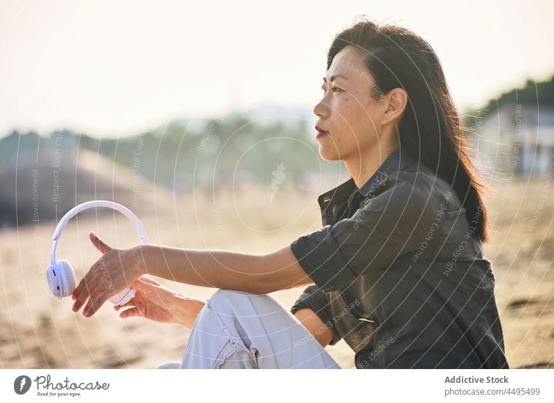 Pensive Asian woman listening to music on beach seaside seashore headphones meloman song pastime thoughtful pensive playlist sand asian female summer recreation