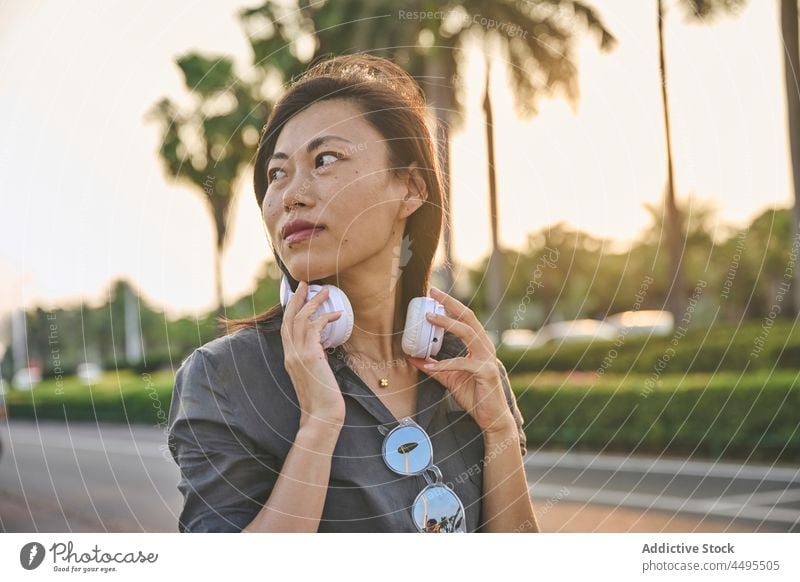 Asian woman with headphones on street meloman style music hobby road town pastime female trendy calm black hair attractive asian summer plant roadway vegetation