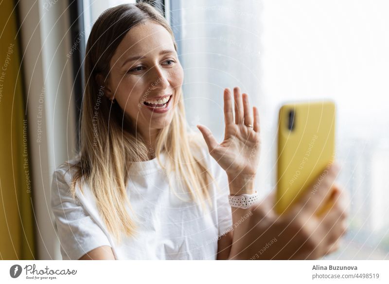 Young pretty millennial woman holding mobile phone at home or cafe near window. video chat food delivery online shopping social media streaming influencer