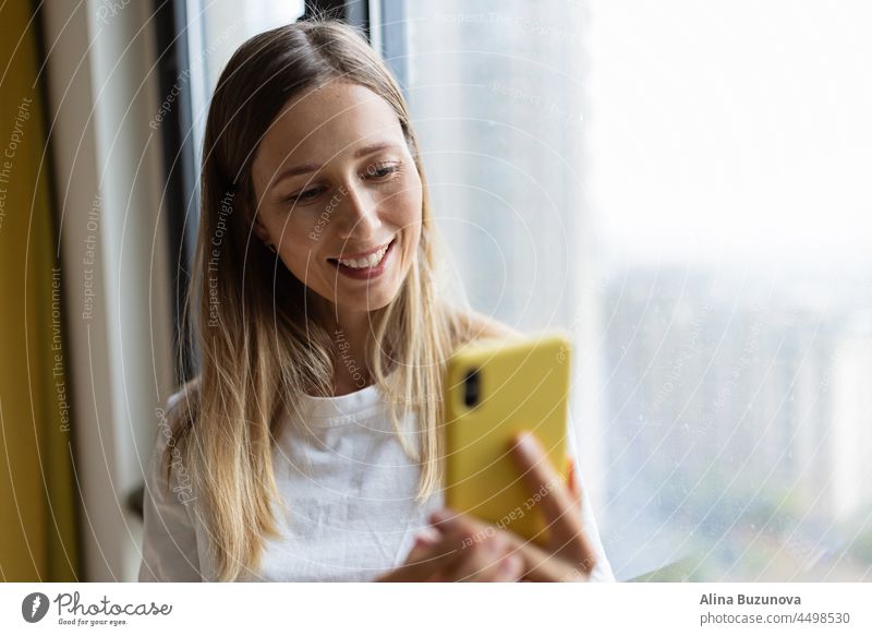 Young pretty millennial woman holding mobile phone at home or cafe near window. video chat food delivery online shopping social media streaming influencer