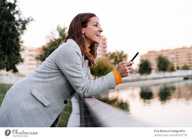 smiling caucasian woman wearing yellow pullover and grey coat using mobile phone device outdoors in park during sunset. Technology and lifestyle autumn speaking