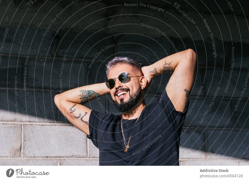 Cheerful male with tattoos standing with raised arms in sunlight man style smile arms raised cheerful positive appearance individuality happy sunglasses trendy