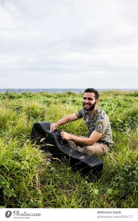 Man musician opening case with guitar on grass in daylight man acoustic instrument hobby shore guitarist male sit calm melody ocean sea nature water song sound