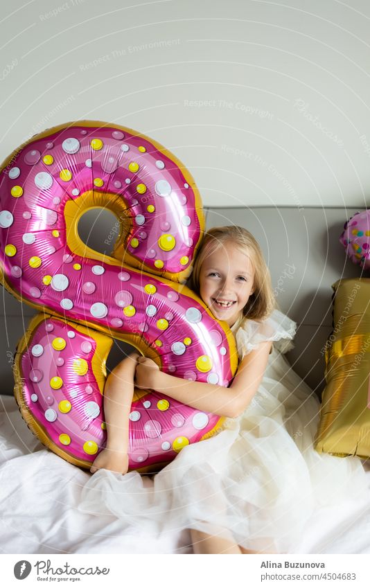 Indoor shot of pretty joyful cute adorable girl kid celebrating eight years old birthday with bright and colorful balloons with inscription words stay fabulous, wearing casual fashionable dress