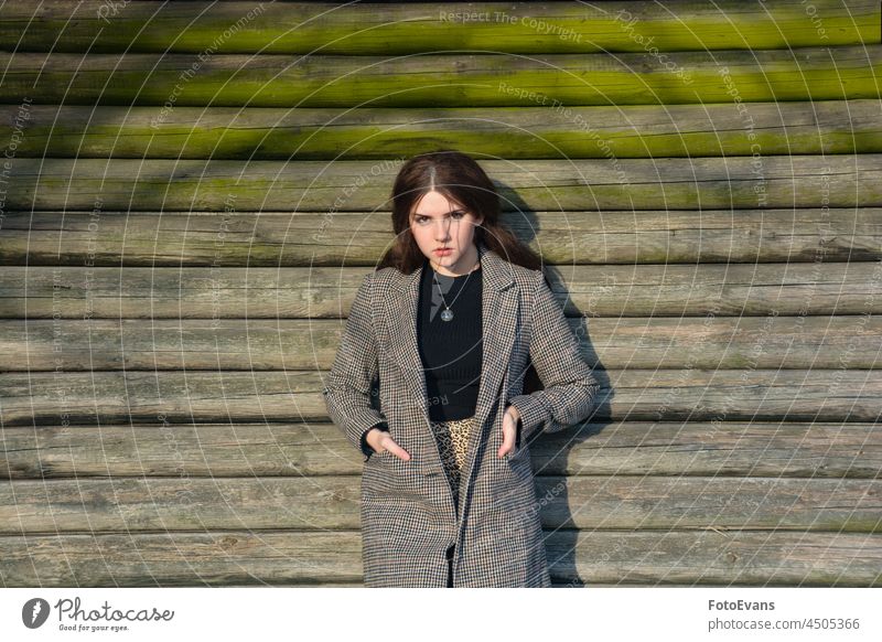 A young girl stands in front of wooden planks sunny fashion brown jacket female outside real coat outdoors sunlight real person bar wooden hut teenager winter