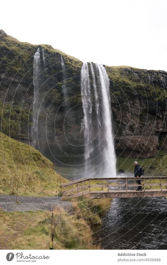 Anonymous man observing waterfall streaming from rocky cliff in Iceland traveler footbridge admire ravine nature explore pond cascade landscape male