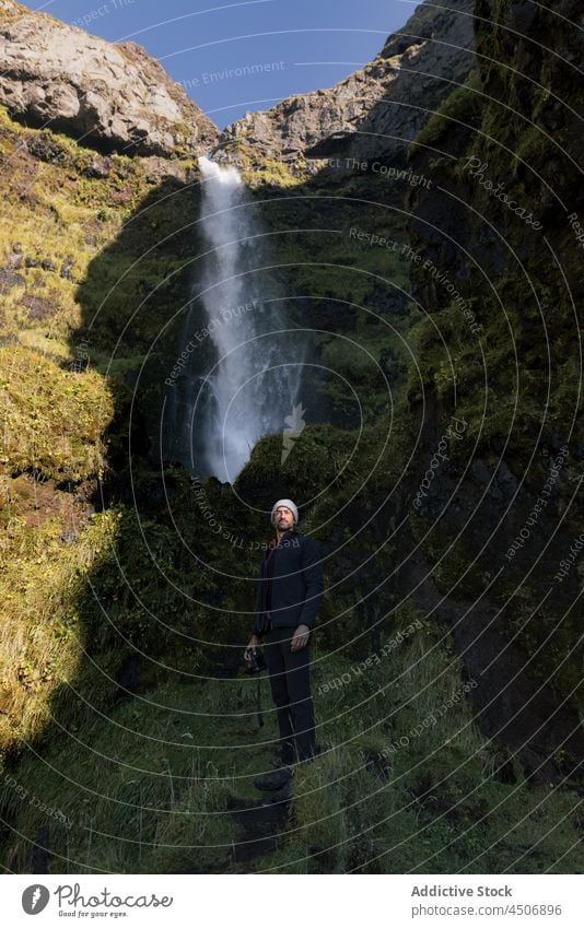 Young ethnic male photographer admiring nature near waterfall during trip in Iceland man admire ravine photo camera traveler explore hike picturesque vacation