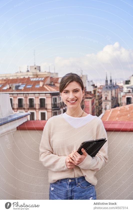 Smiling woman with tablet standing on balcony using internet appearance rooftop positive online charming female phone terrace casual jeans optimist