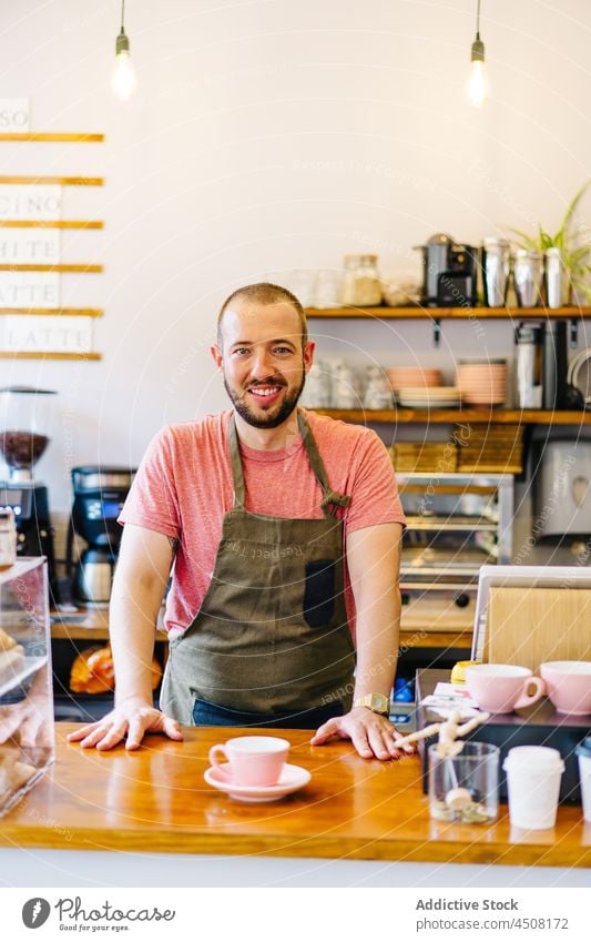 Positive barista standing behind counter man coffee house cup work service staff hot drink professional cafeteria apron male job employee occupation workday