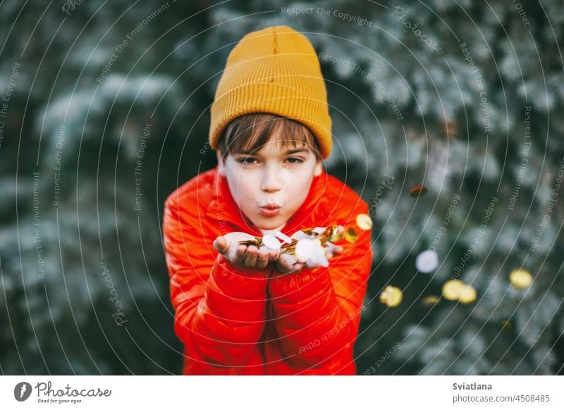 A boy in an orange jacket and hat holds shiny confetti in his hands and blows on it, the boy rejoices at the holidays. Holiday atmosphere, Christmas, New Year