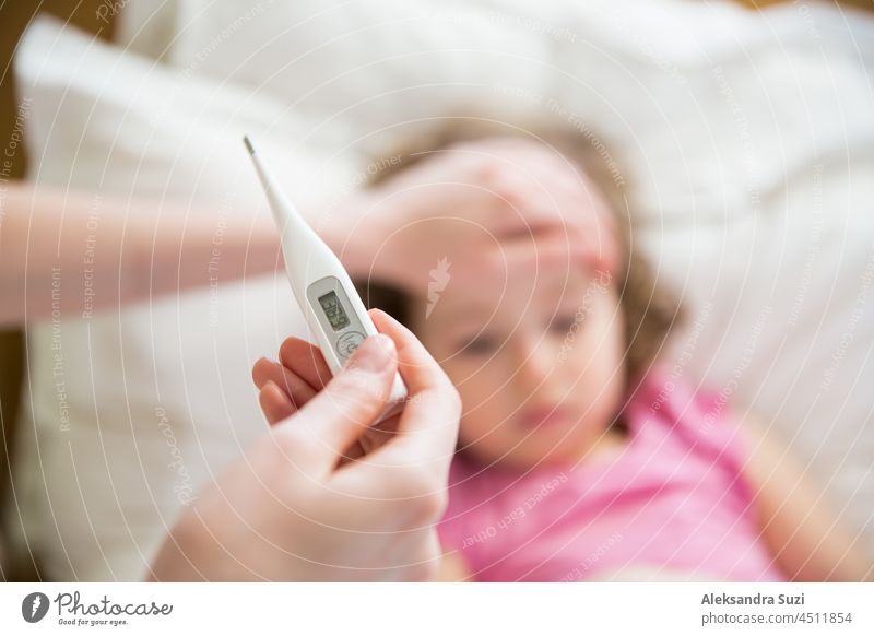 Close-up thermometer. Mother measuring temperature of her ill kid. Sick child with high fever laying in bed and mother holding thermometer. Hand on forehead.