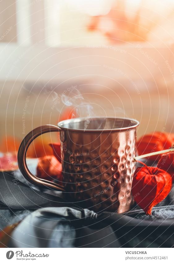 Copper mug with steaming tea on grey wool with orange autumn leaves and physalis at window background with natural light. Cozy autumn time at home with warm drink. Front view.