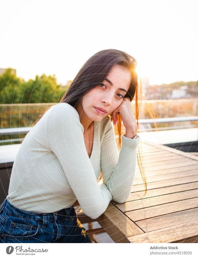 Serious woman sitting at wooden table on street in sunny day joy serious sunlight city female brown hair style casual trendy content glad urban lady fun delight