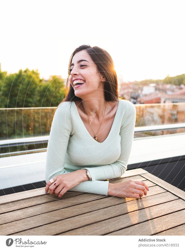 Happy woman sitting at wooden table on street in sunny day laugh cheerful happy joy sunlight city smile female brown hair optimist positive style casual trendy