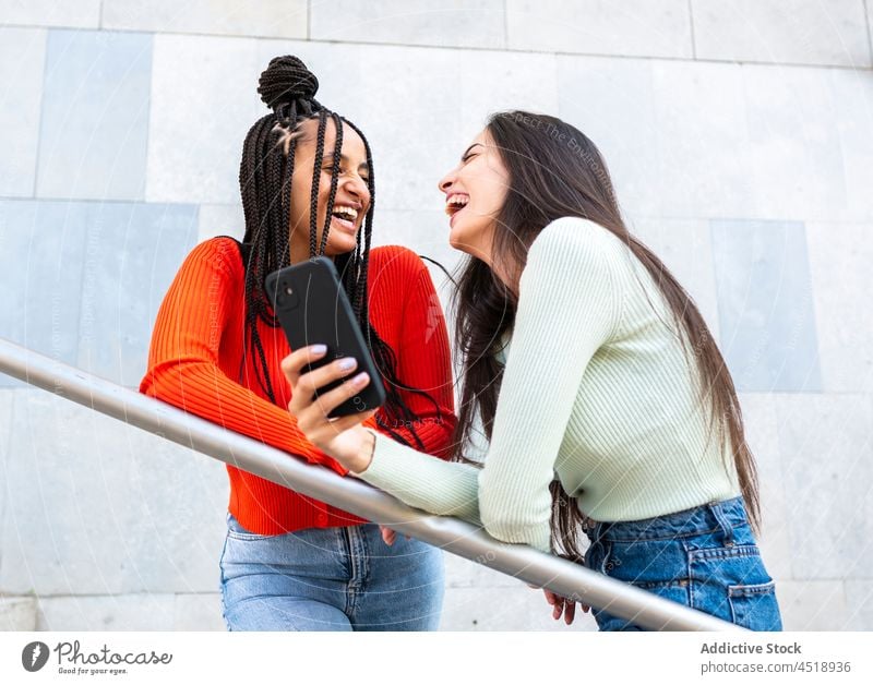 Happy multiracial women friends standing with smartphone near metal railing laugh browsing using cheerful street style female trendy gadget device surfing