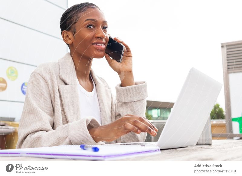 Black woman talking on smartphone near laptop phone call conversation discuss street online freelance work netbook gadget modern computer portable internet