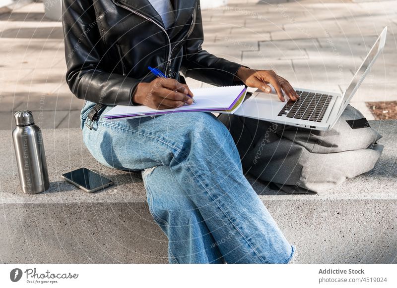 Anonymous black woman browsing laptop while writing in notebook street online internet write netbook information gadget modern computer portable