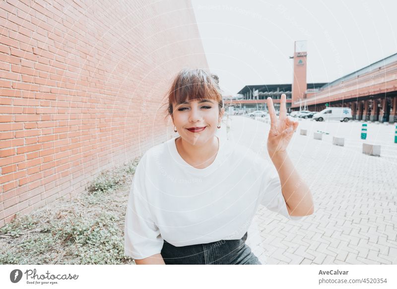 Young redhead hipster girl on white t shirt doing the peace sign while smiling, city lifestyle, young life, black space, sunny day positive selfie mobile