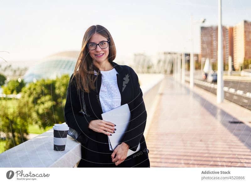 Smiling businesswoman with laptop and takeaway coffee on bridge positive style well dressed formal appearance female to go netbook gadget cup toothy smile