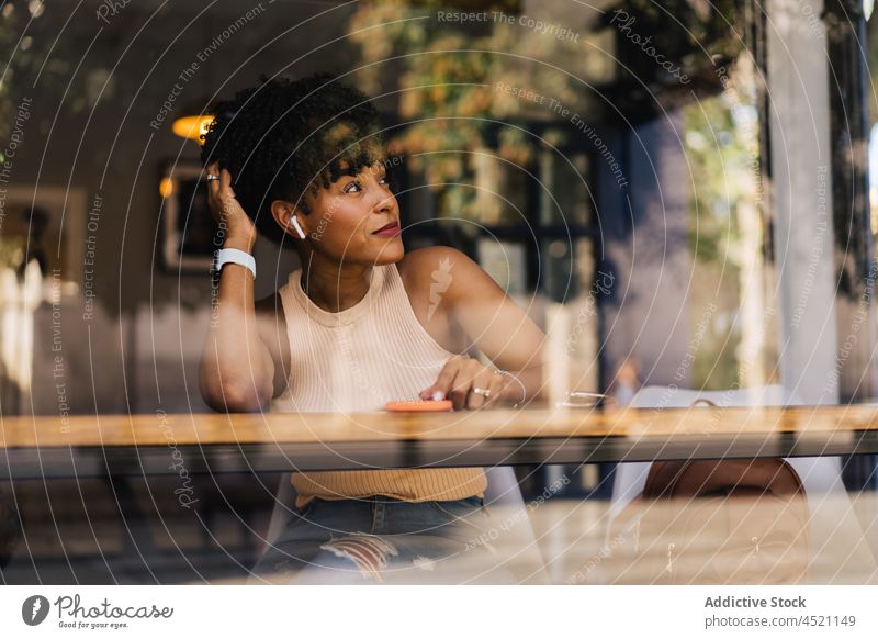 Confident young ethnic lady looking away while resting in cafe woman touch hair trendy dreamy smartphone free time confident window connection style female