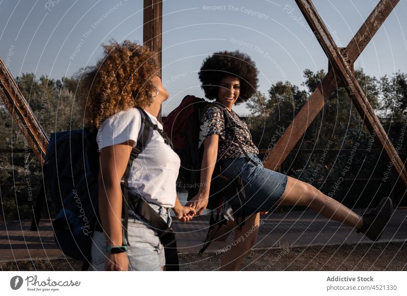 Black friends holding hands on old bridge women laugh joy hike happy walk fun female cuddle best friend carefree adventure raised leg backpack friendship