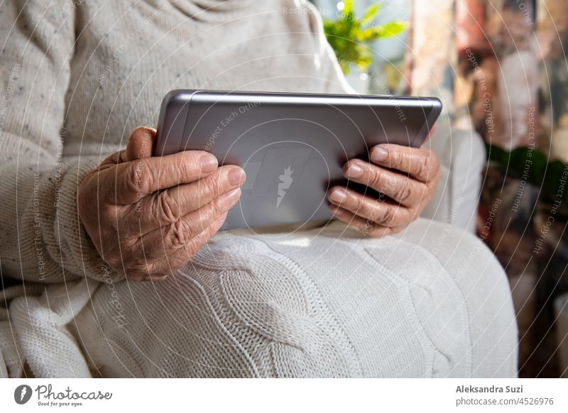 Close-up wrinkled hands of old woman with tablet computer. Senior lady sitting in chair with blanket. Warm and cozy, time to relax, browsing, watching movie.