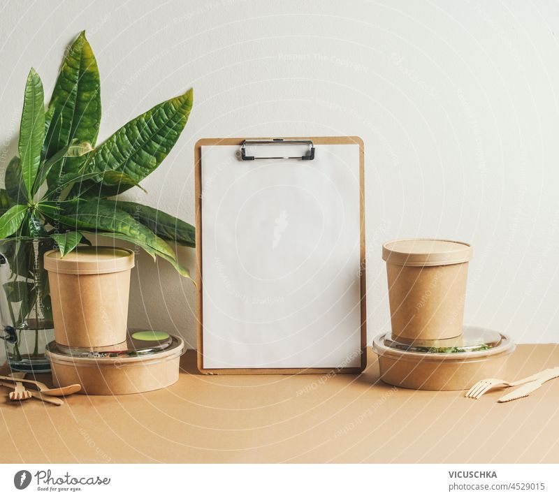 Empty white note board, delivery food in sustainable food containers and leaves on brown table at white wall background. Modern office still life with lunch. Front view with copy space.