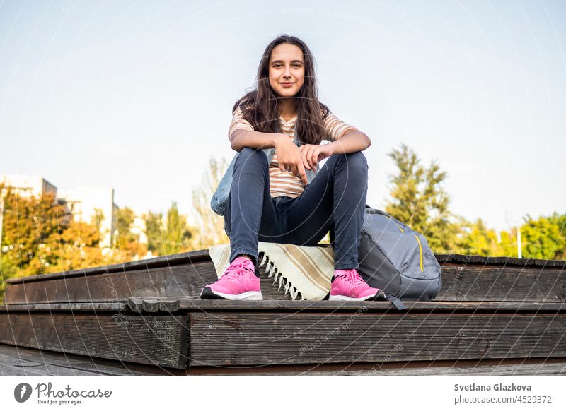 Beautiful and cheerful teen student girl with long brown hair, striped long sleeve shirt and denim vest with backpack sitting in university park on autumn day School pupil Back to school Copy space