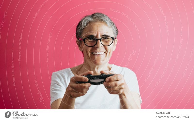 Old senior woman smiling to camera while holding a console pad, video games playing pastel pink removable background, video games old people, copy space, white shirt space