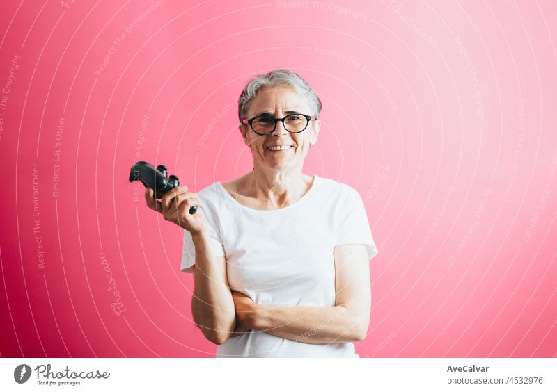 Old senior woman smiling to camera while holding a console pad, video games playing pastel pink removable background, video games old people, copy space, white shirt space