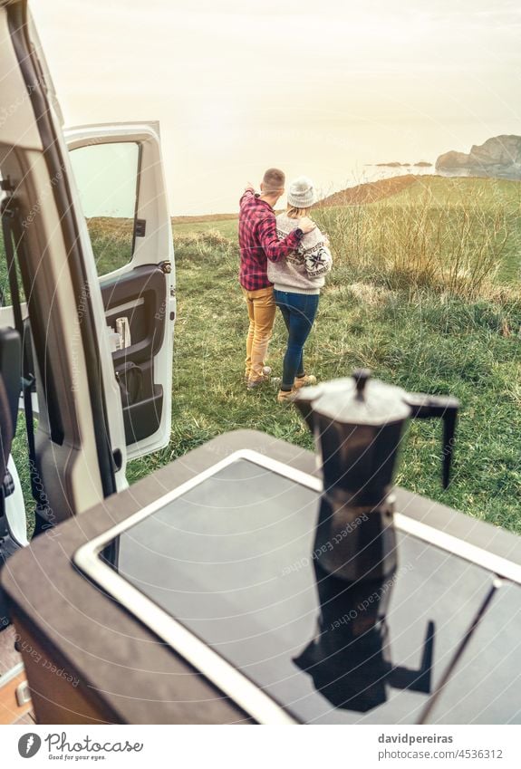 Unrecognizable couple traveling in camper van looking coastline landscape unrecognizable pointing sea morning dawn sunlight coffee cup copy space romantic