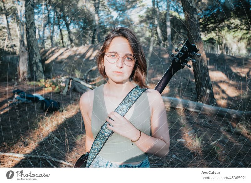 Portrait of an smiling young hipster woman holding and playing the guitar outside the forest park of the city. Having fun learning a new skill, music play seasonal style. Copy space