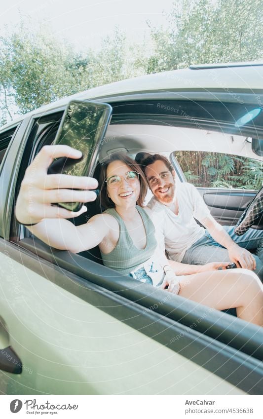 Lovely young couple traveling doing a road trip in car. Road travel, frontal portrait smiling. They are taking a break from driving and taking a selfie with their phone. Hipster modern couple.