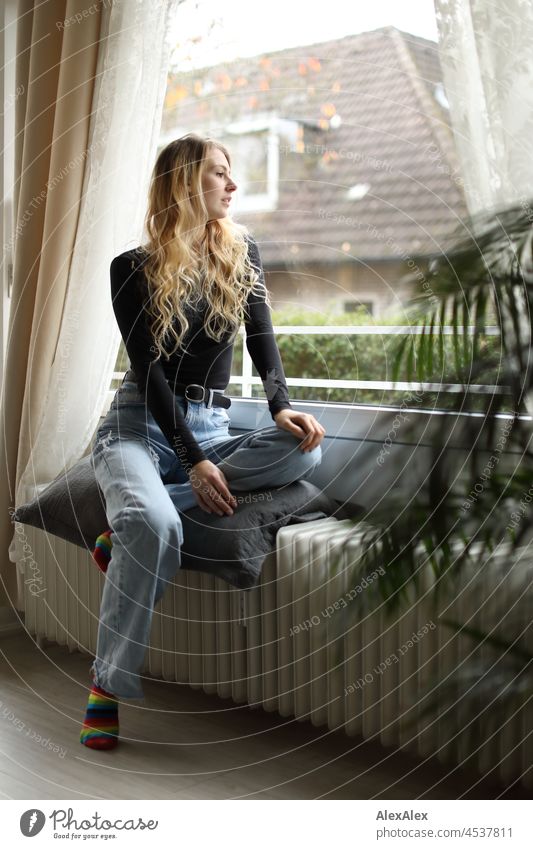 Young, slim, tall woman with long blond hair sits in the living room in front of the balcony window on the heater Woman Young woman Large Blonde Long-haired