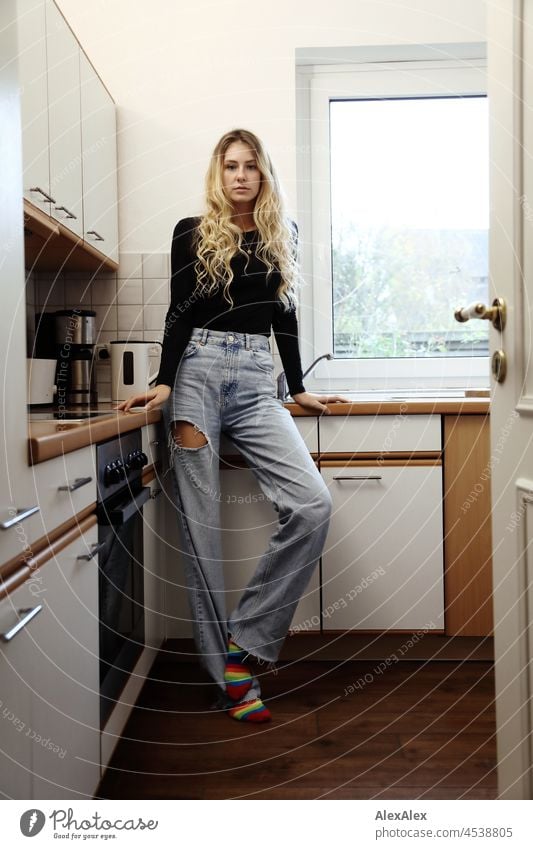 Young, slim, tall woman with long, blond hair stands in a kitchen leaning against the cupboard and looks into the camera Woman Young woman Large Blonde