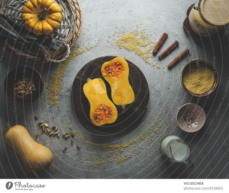 Halved butternut squash on plate with bowl of millet and spices: cardamom, cinnamon on grey concrete kitchen table. Preparing healthy vegan food with seasonal ingredients at home. Top view