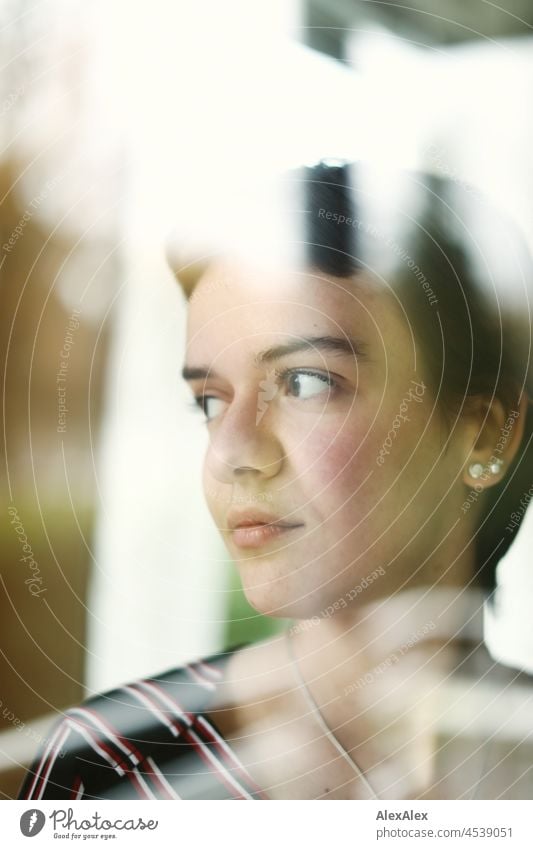 Young woman with short brunette hair looks out the window behind the glass Woman short hair Brunette pretty naturally Authentic Jewellery Chain Earring Slim