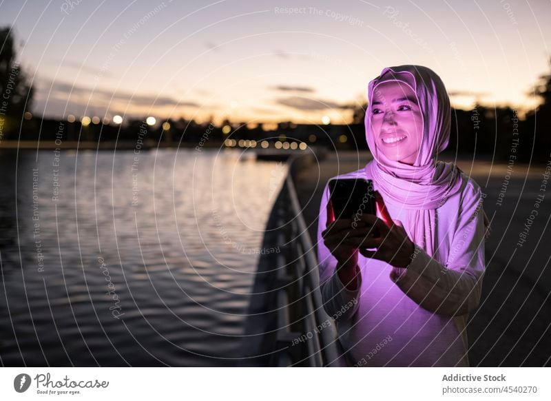 Positive Muslim woman using smartphone on embankment purple light sundown positive dark connection female tradition hijab sunset online mobile smile glow