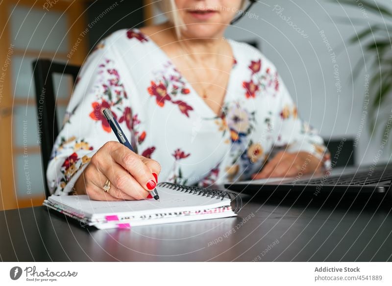 Anonymous mature woman writing in notebook while working on laptop write using concentrate busy remote job female take note self employed telework device