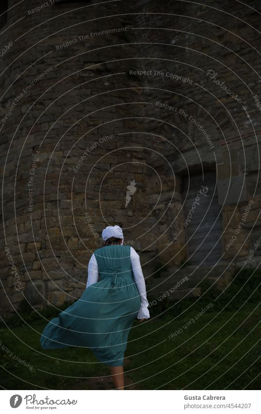 Woman in Victorian dress, walks towards a stone house with a wooden door. Victorian style victorian victorian era Colour photo Exterior shot Feminine Dress