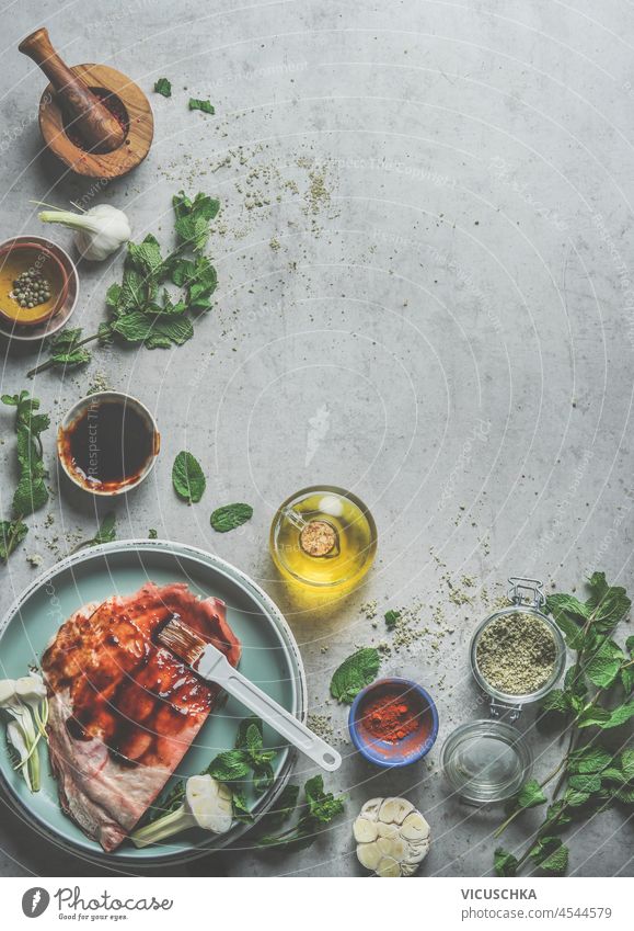 Food background with raw meat, steak marinade, spices, oil, mortar and pestle, mint and garlic on grey concrete kitchen table. BBQ or grill preparation at home with flavorful ingredients. Top view.