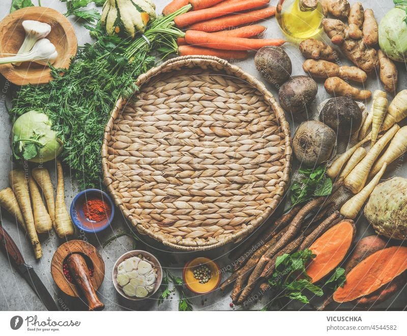 Various raw root vegetables around plaited kitchen tray on grey table. Autumn vegetables: beetroot, parsnip, sweet potato, kohlrabi, celery, garlic, salsify. Top view. Frame with copy space.