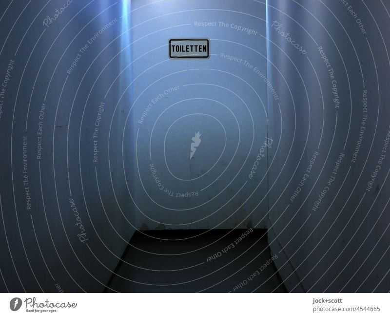 a bluish corridor leads to the TOILETS Corridor Artificial light Blue Signs and labeling toilets Signage Word Monochrome Wall (building) Neutral Background