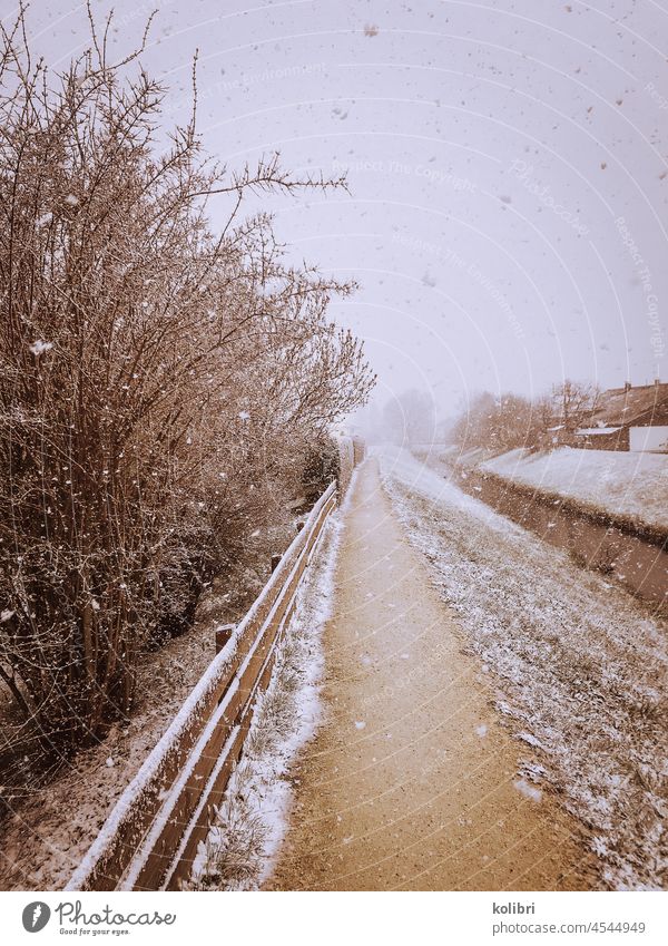 Footpath in winter, always straight ahead, on the left of it a wooden fence, behind it bushes, on the right side of the path a brook bank. Falling snowflakes, in the distance trees can be seen in the mist.