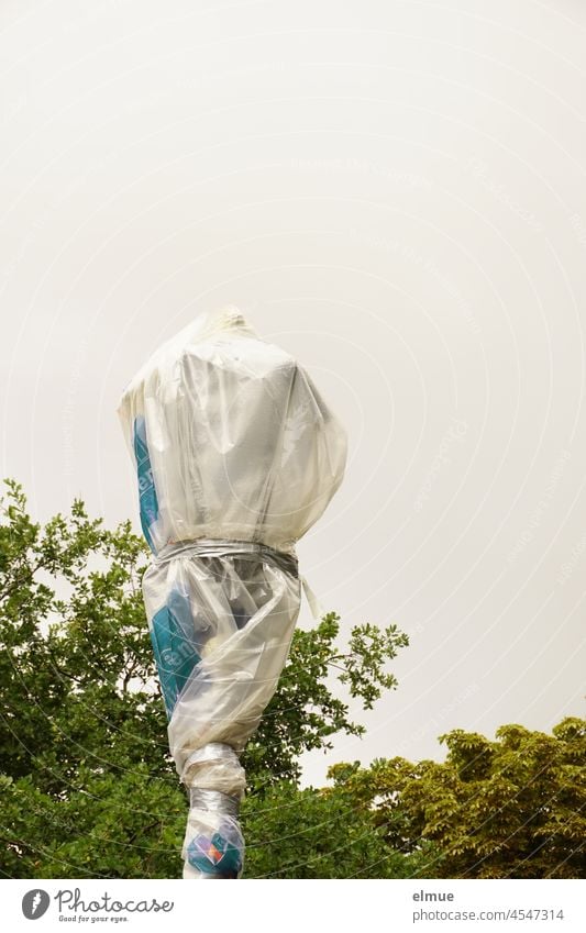 a street lamp still wrapped in foil stands in front of green trees Packaged New Street lighting streetlamp Lantern Lamp post Lighting shrouded Packing film